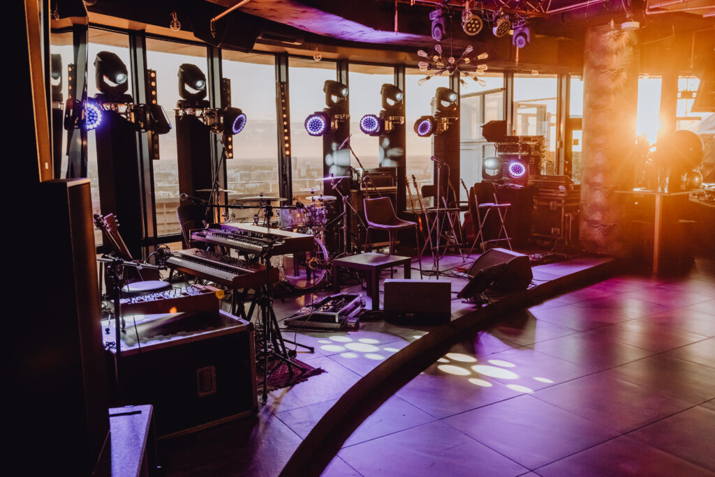 Na zdjęciu widoczna jest profesjonalnie przygotowana scena muzyczna w nowoczesnym klubie lub rooftop barze. Znajdują się tam instrumenty muzyczne, mikrofony, perkusja, keyboardy oraz rozbudowane oświetlenie sceniczne. Duże okna w tle pokazują panoramę miasta, a ciepłe światło zachodzącego słońca nadaje wnętrzu klimatyczny charakter.